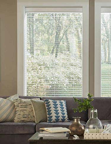 Smart wood blinds in a window behind a blue couch with pillows and gray coffee table in front